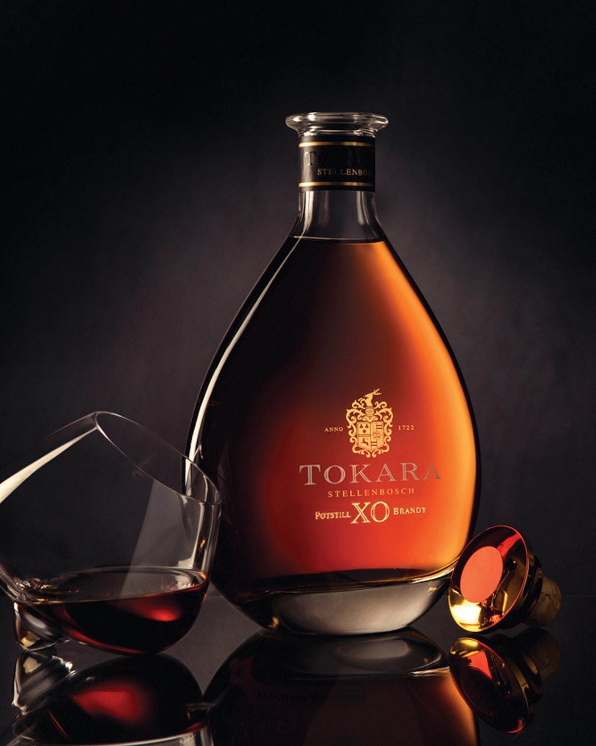 Eine Flasche Tokara Stellenbosch XO Brandy steht aufrecht, daneben steht ein Schnapsglas, und der gold-rote Verschluss der Flasche liegt auf der Seite, alles vor einem dunklen Hintergrund.