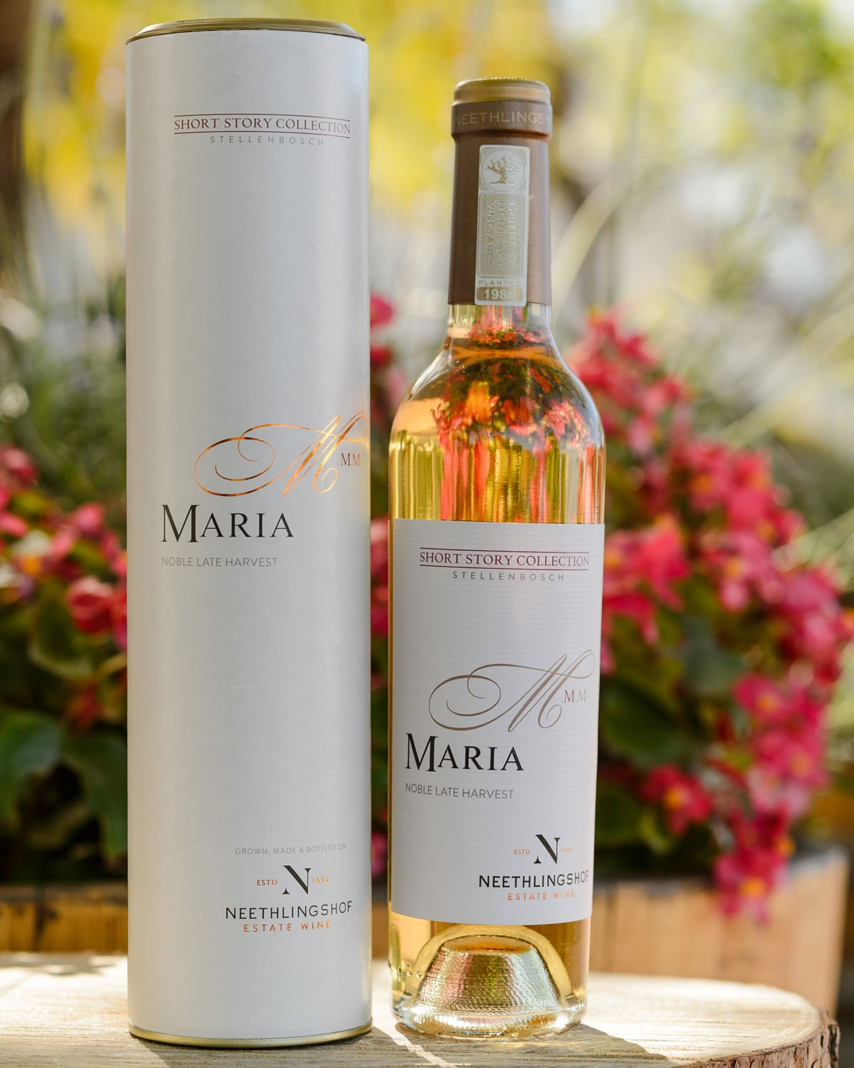 Eine Flasche des Maria Noble Late Harvest Weins vom Weingut Neethlingshof steht neben der dazugehörigen zylindrischen Kiste, während im Hintergrund bunte Blumen und Grünpflanzen verschwimmen.