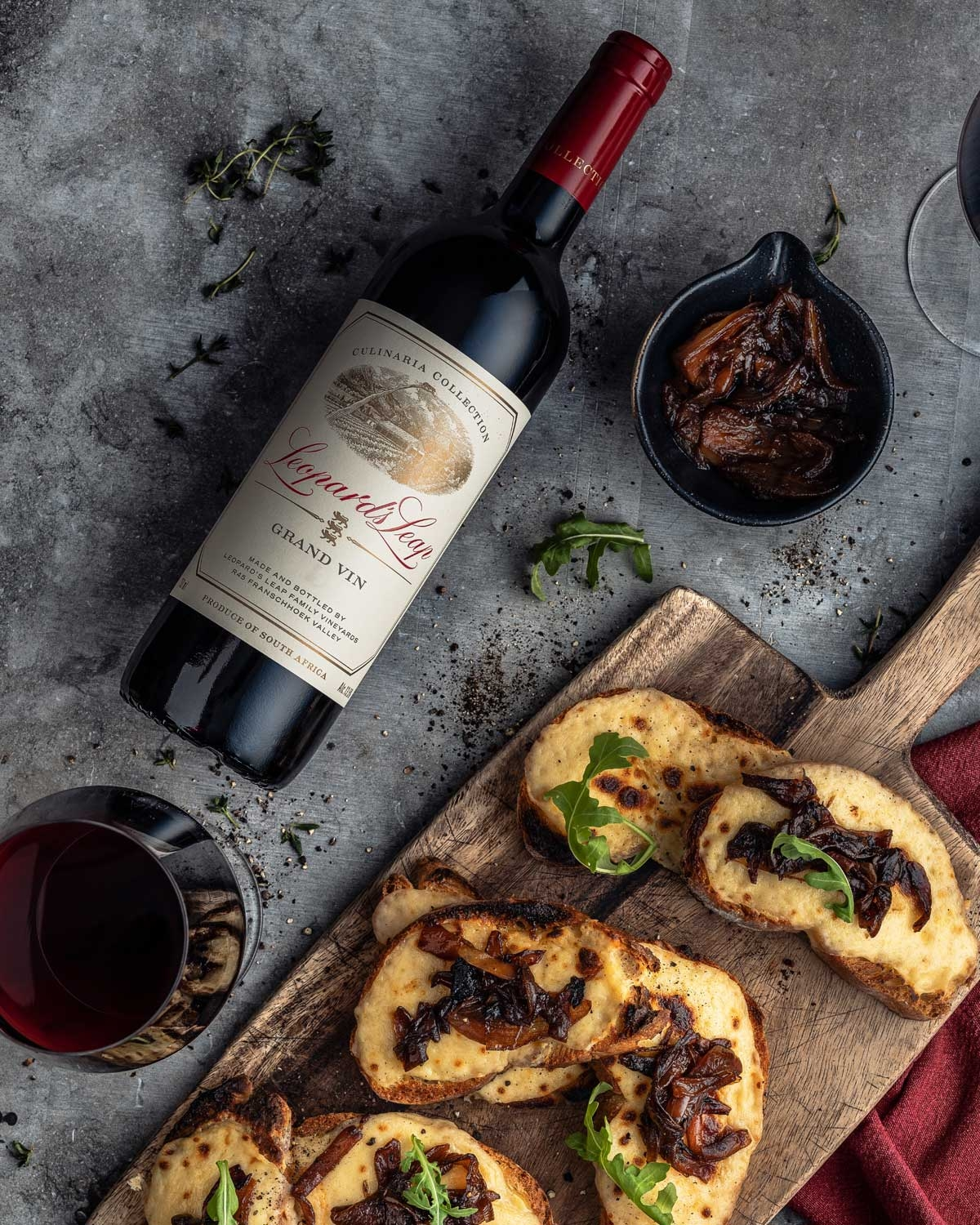 Eine Flasche Rotwein, ein Glas Wein, getoastetes Brot mit karamellisierten Zwiebeln und Rucola auf einem Holzbrett und eine kleine Schale mit karamellisierten Zwiebeln auf einer grauen Fläche mit Kräutern drumherum verstreut.