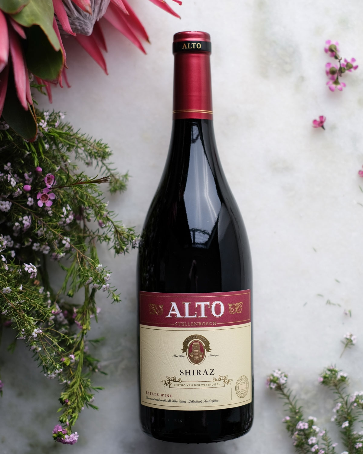 Eine Flasche Alto Shiraz Rotwein liegt auf einer hellen Marmorfläche, umgeben von rosa und weißen Blumen und Grünzeug. Das Weinetikett ist rot und cremefarben mit goldenen Akzenten.
