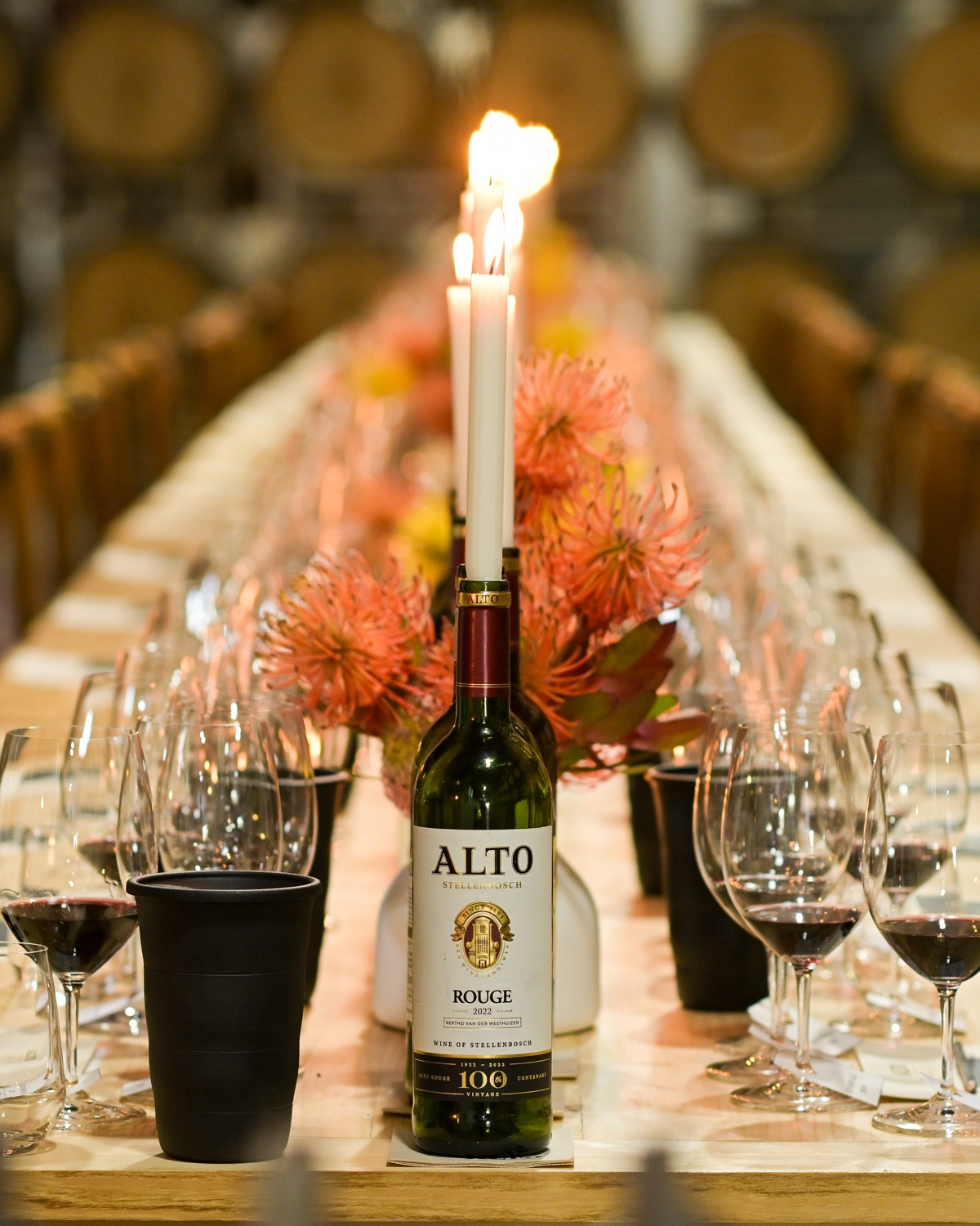 Eine Weinflasche mit zwei hohen brennenden Kerzen steht auf einem langen, elegant gedeckten Esstisch mit Weingläsern, orangefarbenen Blumen und einem verschwommenen Hintergrund aus Weinfässern.