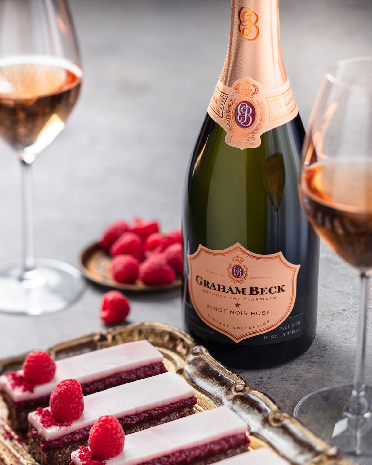 Eine Flasche Graham Beck Pinot Noir Rosé steht neben zwei Gläsern Roséwein, einem kleinen Teller mit frischen Himbeeren und einem Tablett mit Himbeerdesserts, die mit weißer Glasur und Himbeeren belegt sind.