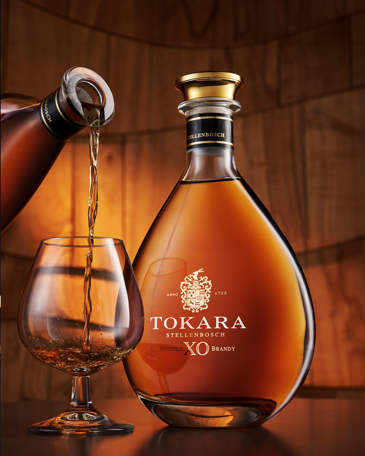 Ein Glas Tokara Stellenbosch XO Brandy wird neben einer großen, eleganten Flasche desselben Brandys vor einem warmen, hölzernen Hintergrund eingeschenkt.