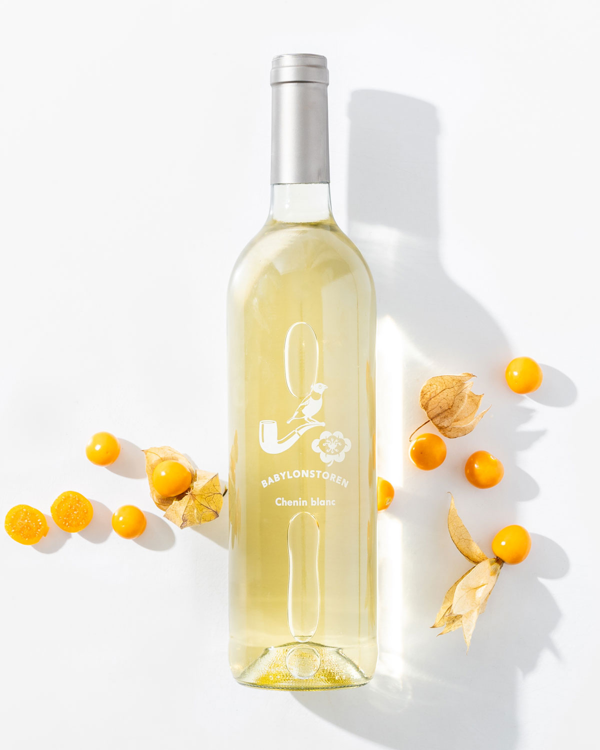 Eine durchsichtige Flasche Babylonstoren Chenin Blanc steht aufrecht, umgeben von gelben Kapstachelbeeren und ihren papierartigen Schalen auf weißem Grund.