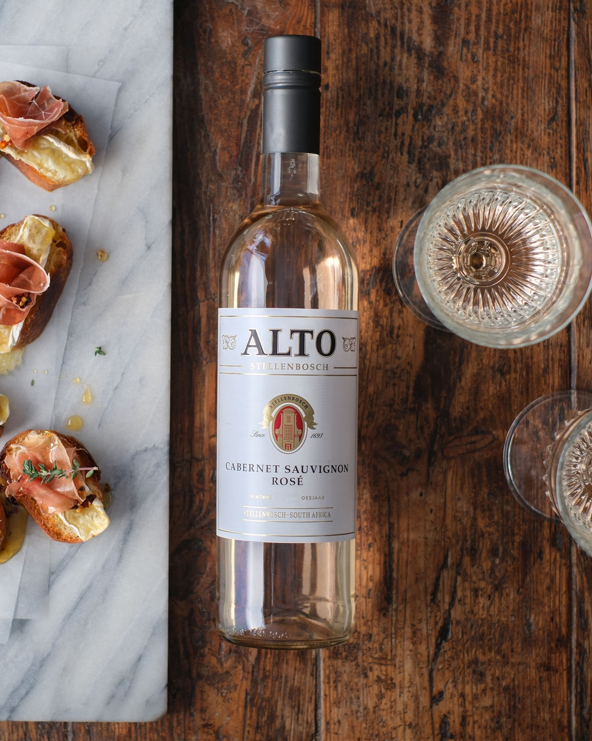 Eine Flasche Alto Cabernet Sauvignon Rosé steht auf einem Holztisch neben zwei gefüllten Weingläsern und einem Marmorbrett mit Bruschetta, garniert mit gepökeltem Fleisch und Kräutern.