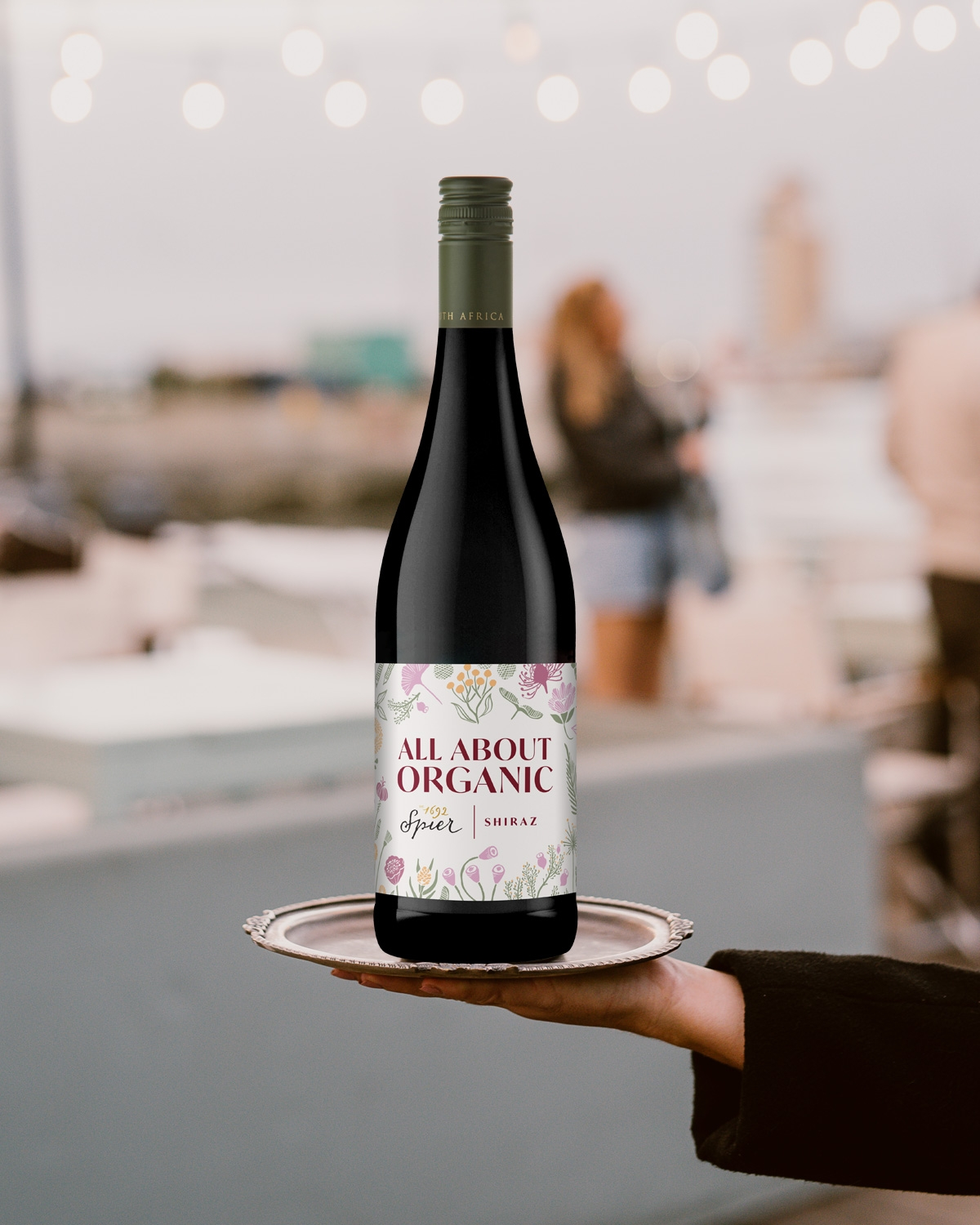 Eine Hand hält ein silbernes Tablett mit einer Flasche "All About Organic" Shiraz-Wein. Im Hintergrund sind Lichterketten und Menschen zu sehen, die eine festliche Atmosphäre im Freien schaffen.
