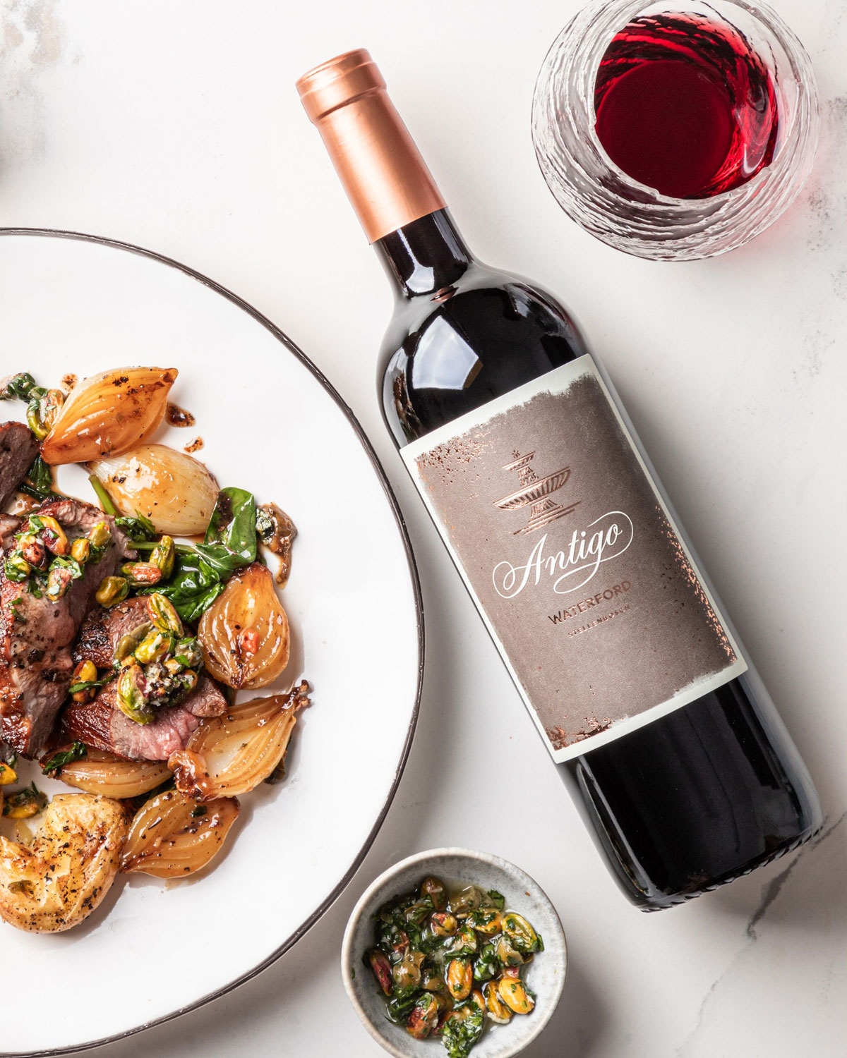 Eine Flasche Antigo-Wein neben einem Glas Rotwein, ein Teller mit gebratenem Fleisch, Zwiebeln und Grünzeug und eine kleine Schale mit Soße zum Garnieren, alles auf einer weißen Fläche.