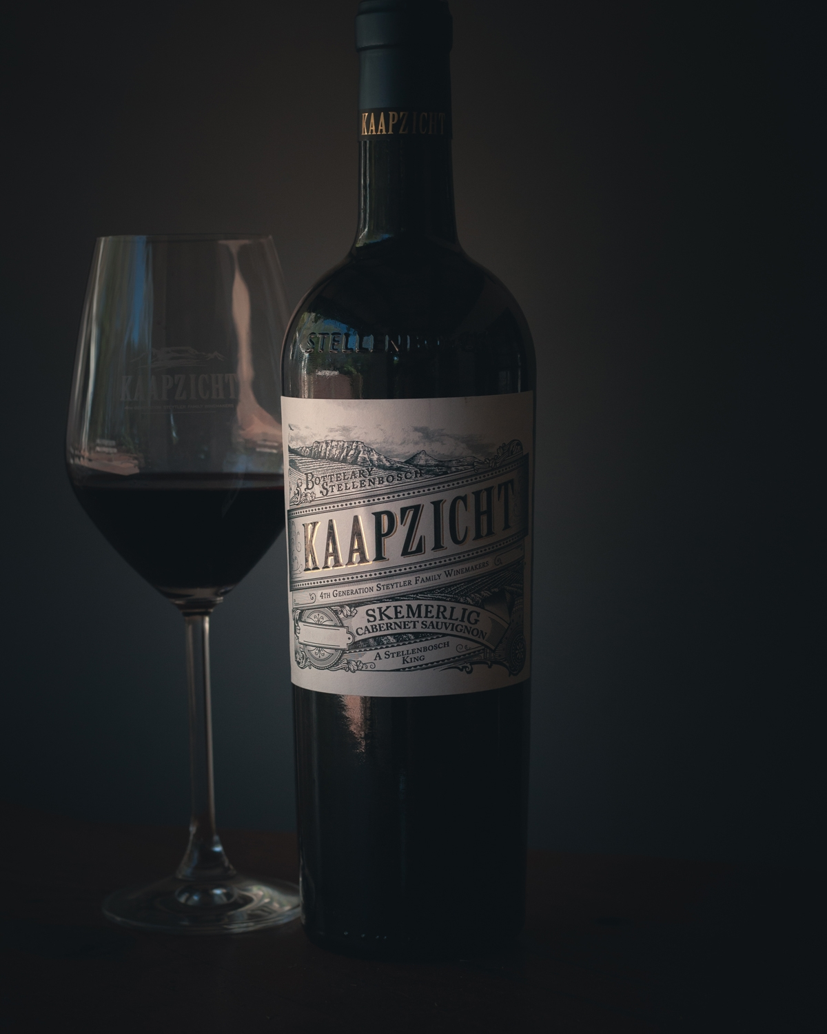 Eine Flasche Kaapzicht Skemerlig Cabernet Sauvignon Rotwein steht neben einem gefüllten Weinglas auf einem dunklen Hintergrund, beide mit dem Kaapzicht-Logo beschriftet.