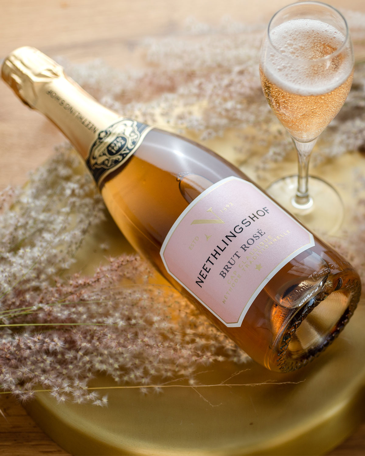 Eine Flasche Neethlingshof Brut Rosé Sekt mit Goldfolie, rosa Etikett und eleganter schwarz-goldener Halsverzierung, mit der Aufschrift "Neethlingshof" und "Brut Rosé Méthode Cap Classique".