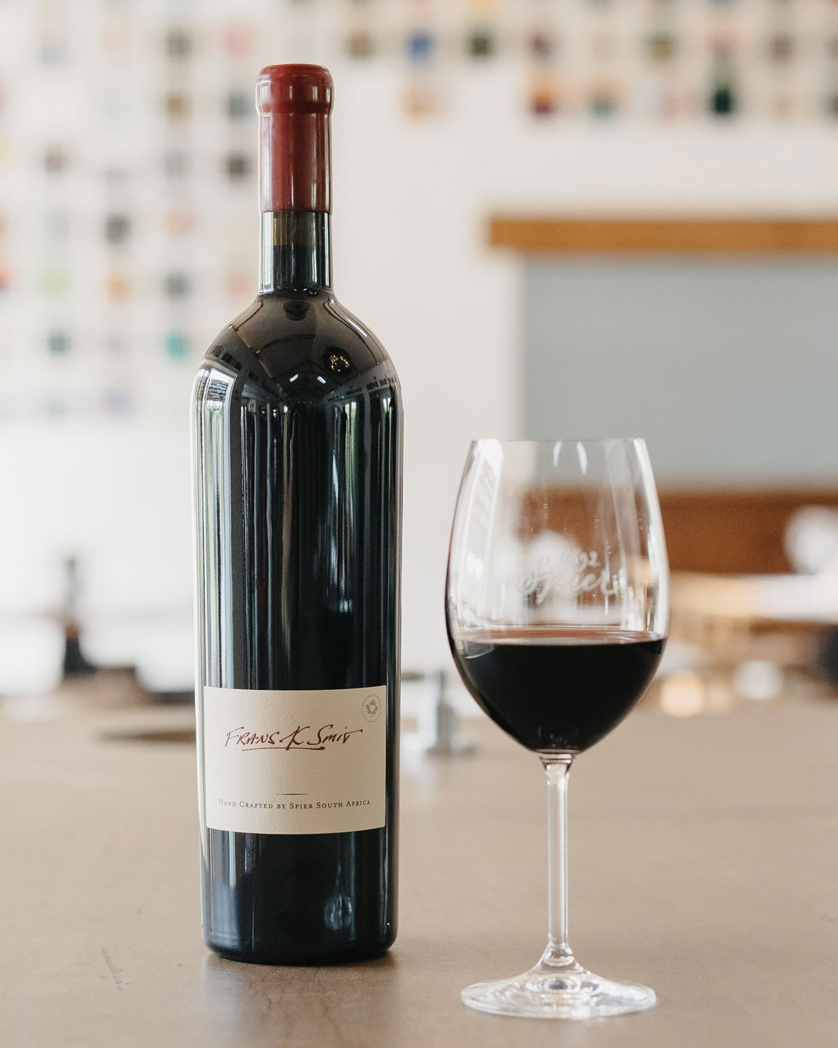 Eine Rotweinflasche mit der Aufschrift "Frans K. Smit" steht neben einem gefüllten Weinglas auf einem Tisch, vor einem weich verschwommenen Hintergrund mit Regalen und bunten Quadraten an der Wand.