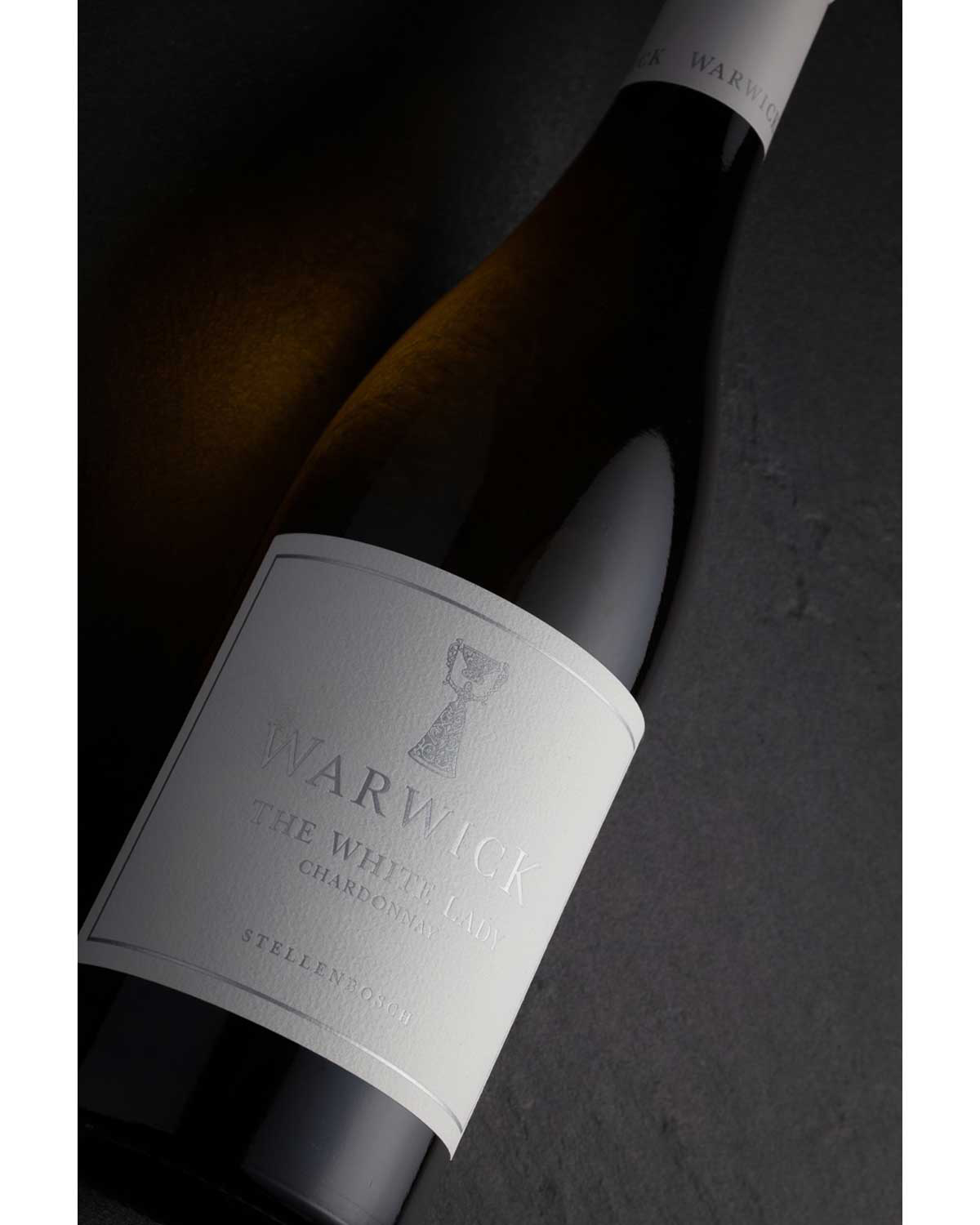 Eine Flasche Warwick The White Lady Chardonnay Wein mit weißem Etikett und Verschluss, mit einfachem silbernem Text und einem Logo vor einem schlichten weißen Hintergrund.