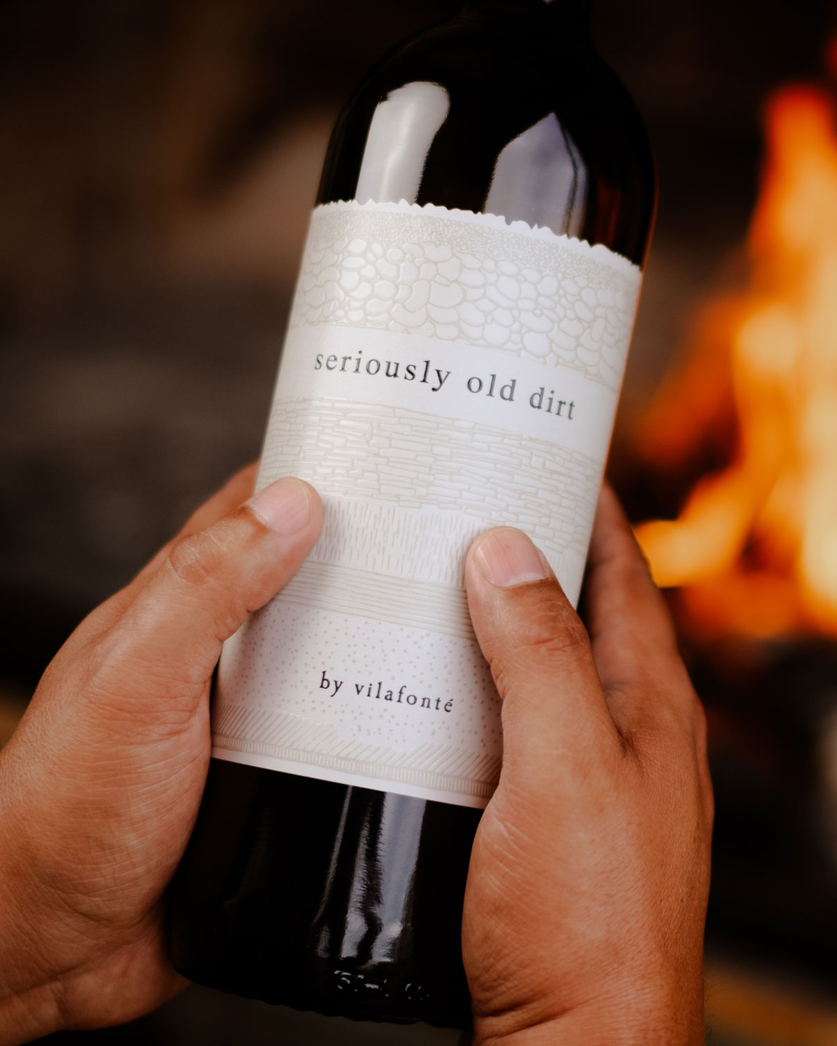 Eine Person hält eine Weinflasche mit der Aufschrift "serious old dirt by vilafonté", während im Hintergrund ein unscharfes Feuer brennt.