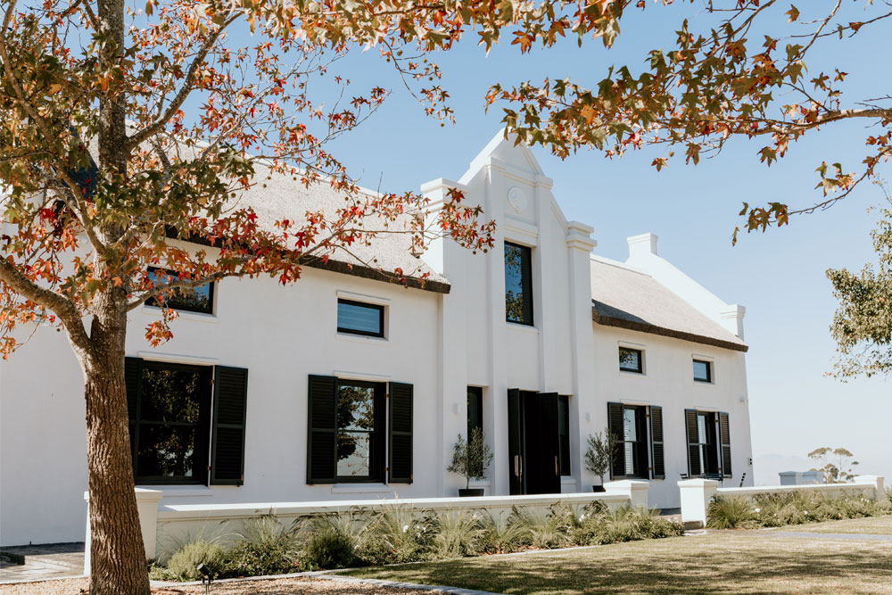 Ein großes weißes Haus mit schwarzen Fensterläden und einem Giebeldach steht hinter einem Baum mit Herbstlaub. Der Himmel ist klar und blau, und der Rasen und die Sträucher sind ordentlich gepflegt.