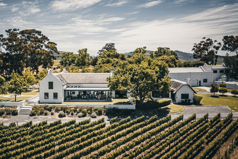Luftaufnahme eines weißen Bauernhauses, umgeben von üppigen Weinbergen und Bäumen unter einem teilweise bewölkten Himmel, mit grünen Hügeln im Hintergrund.