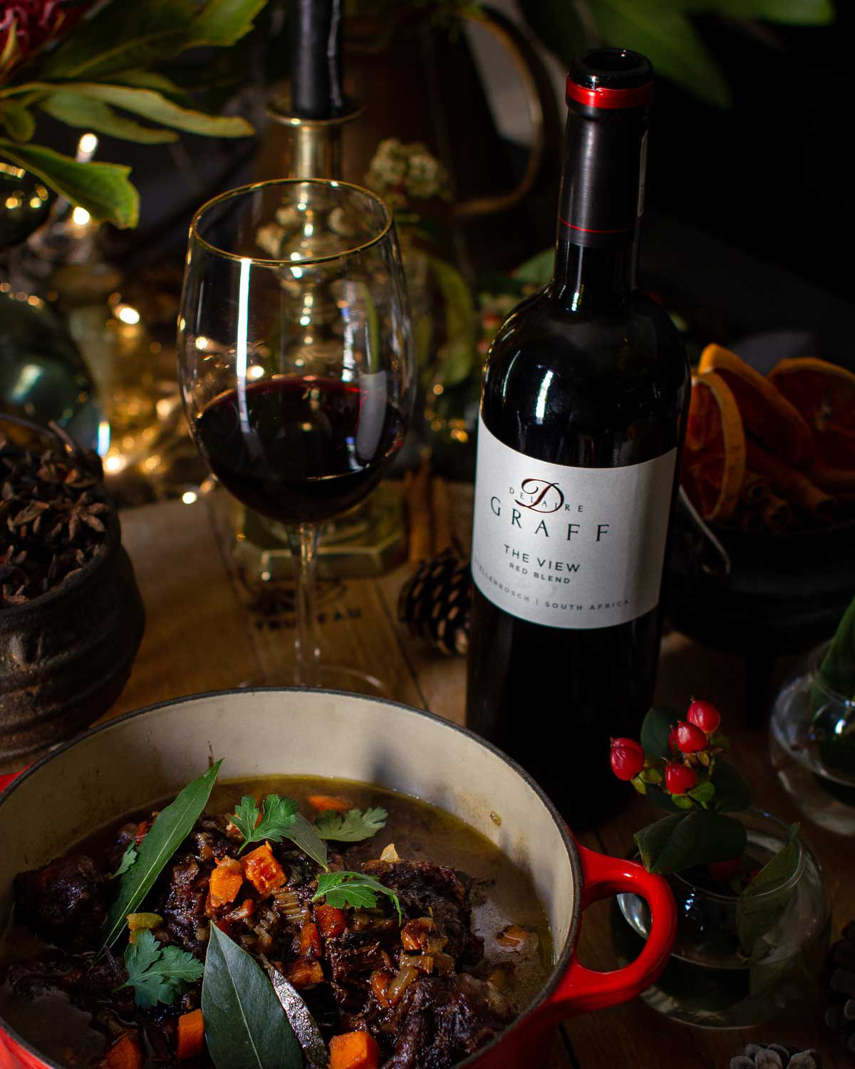 Ein rustikales Gericht aus geschmortem Fleisch mit Karotten und Kräutern wird in einem roten Topf neben einem Glas und einer Flasche Rotwein auf einem Holztisch serviert, umgeben von Blumen und getrockneten Orangenscheiben.