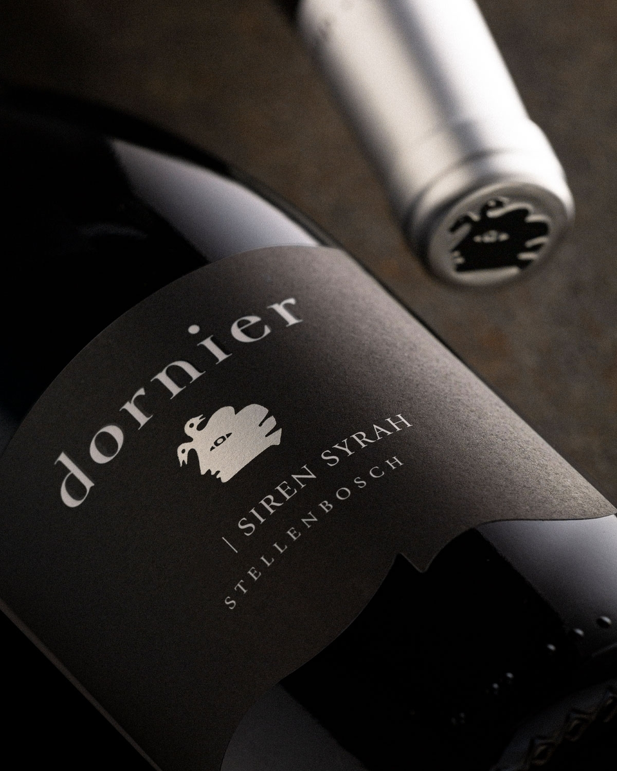Nahaufnahme einer Flasche Dornier Siren Syrah Wein aus Stellenbosch, mit einem schwarzen Etikett mit stilisiertem Logo und silberner Schrift. Die Flasche liegt auf der Seite neben einem passenden Weinverschluss.