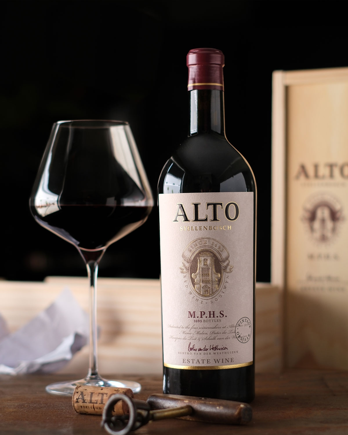Eine Flasche Alto-Wein steht neben einem gefüllten Weinglas auf einer hölzernen Oberfläche, mit einem Korken, einem Korkenzieher und einer hölzernen Weinkiste im Hintergrund. Die Szene ist sanft beleuchtet und hebt das Etikett des Weins hervor.