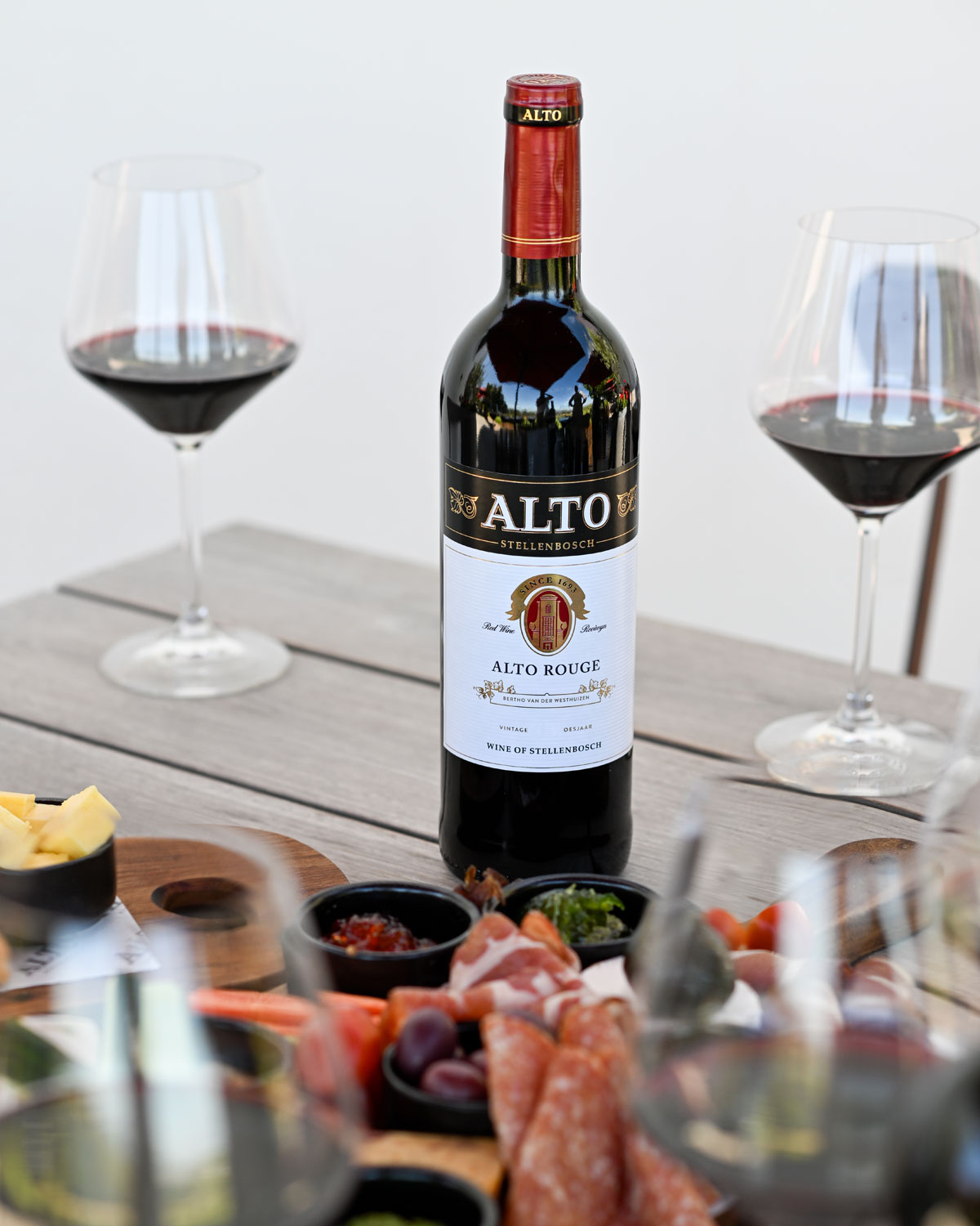 Eine Flasche Alto Stellenbosch Rouge Rotwein mit einer schwarz-goldenen Folienkapsel. Auf dem Etikett finden sich goldene und schwarze Akzente, das Alto-Logo und der Hinweis auf den 100. Jahrgang und den Wein von Stellenbosch.