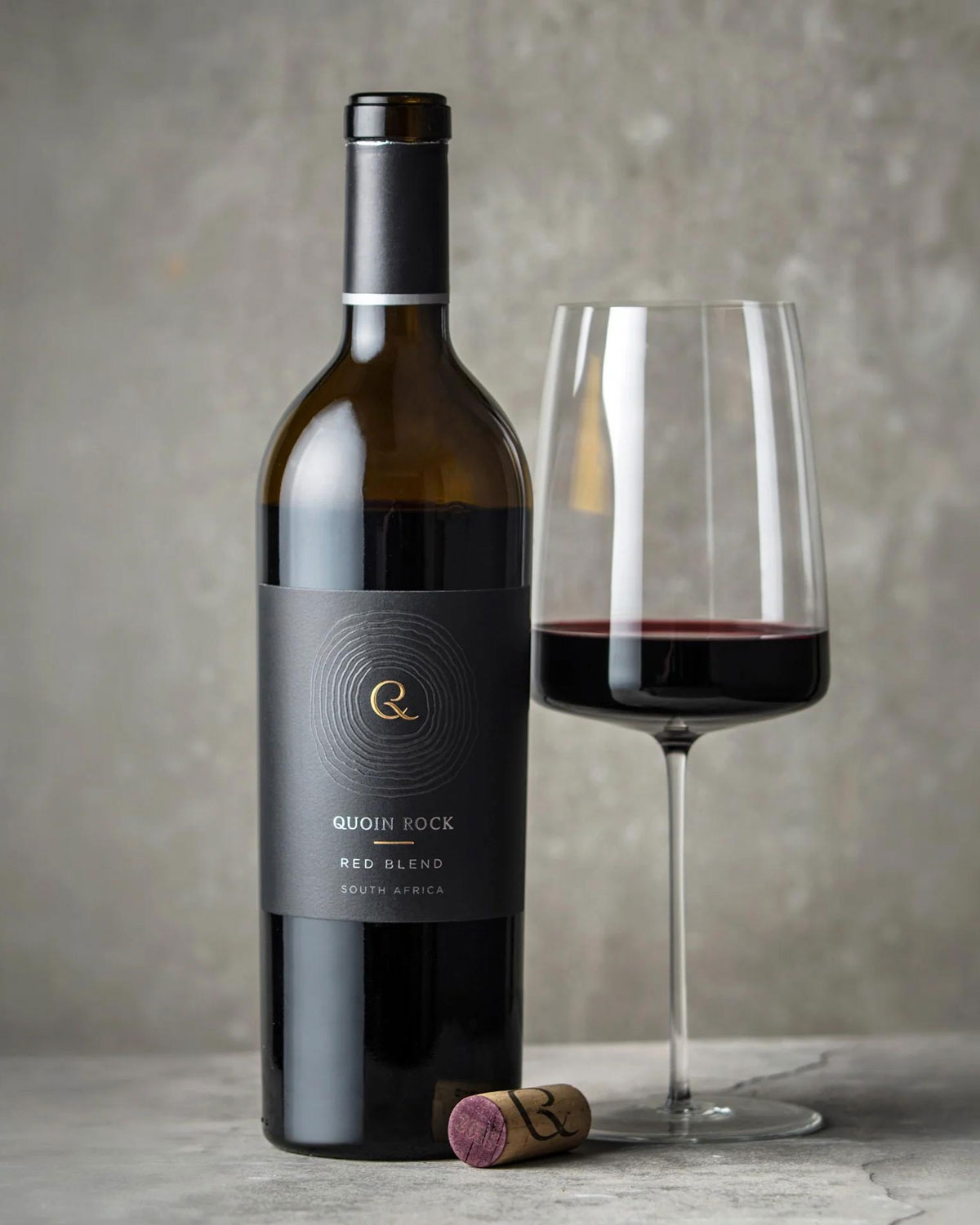 Eine Flasche Quoin Rock Red Blend Wein aus Südafrika steht neben einem gefüllten Weinglas und einem Korken vor einem neutralgrauen Hintergrund.