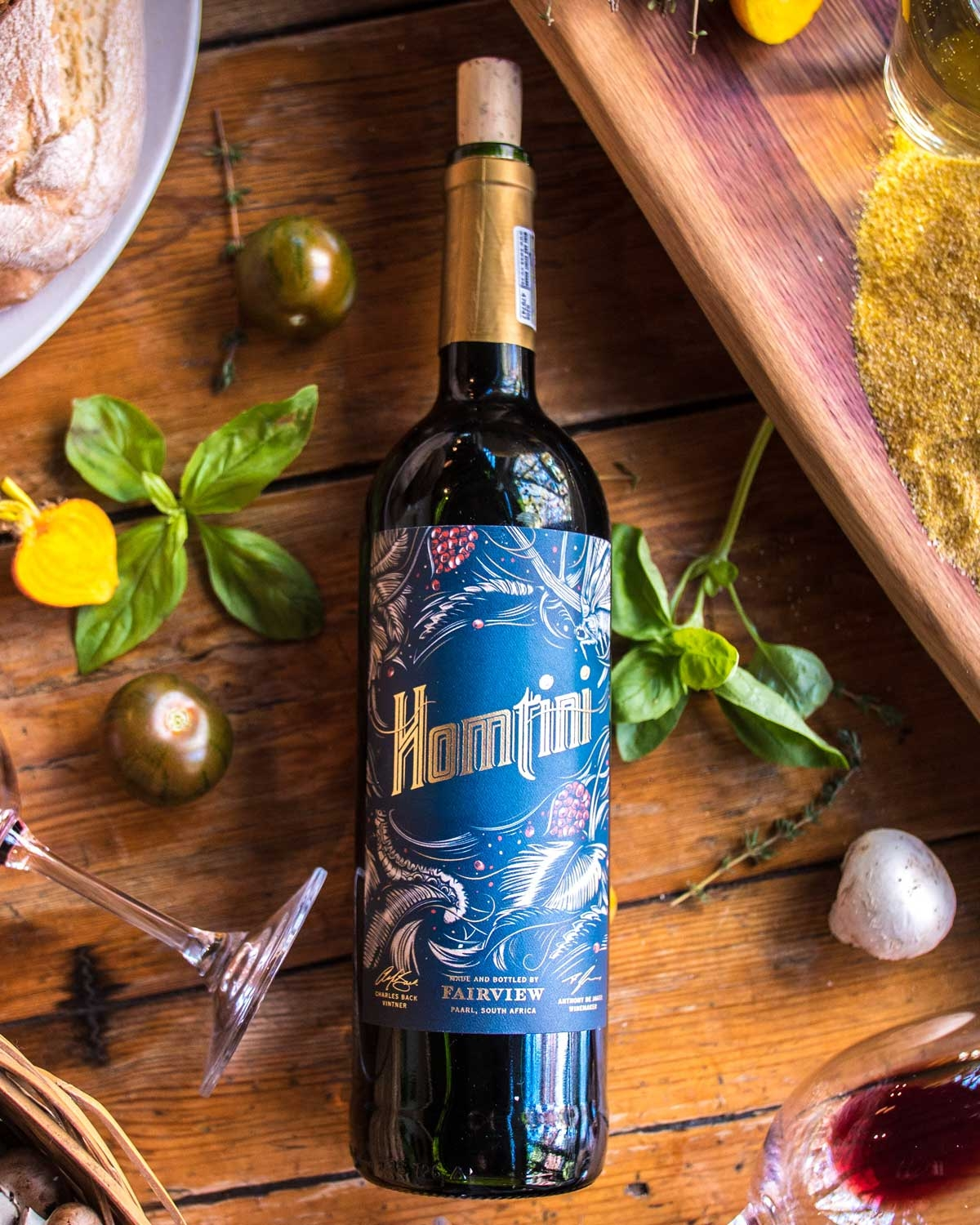 Eine Weinflasche aus dunklem Glas mit goldenem Verschluss und blauem Etikett, das mit Abbildungen von Blättern und Trauben verziert ist. Auf dem Etikett steht in großen, verschnörkelten Lettern "Homtini".