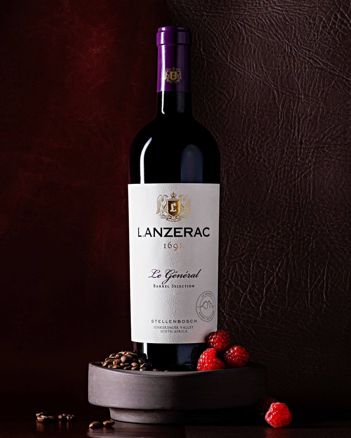 Eine Flasche Lanzerac-Rotwein steht aufrecht auf einem runden Steinpodest, umgeben von frischen Himbeeren und Kaffeebohnen, vor einem dunkel strukturierten Hintergrund.