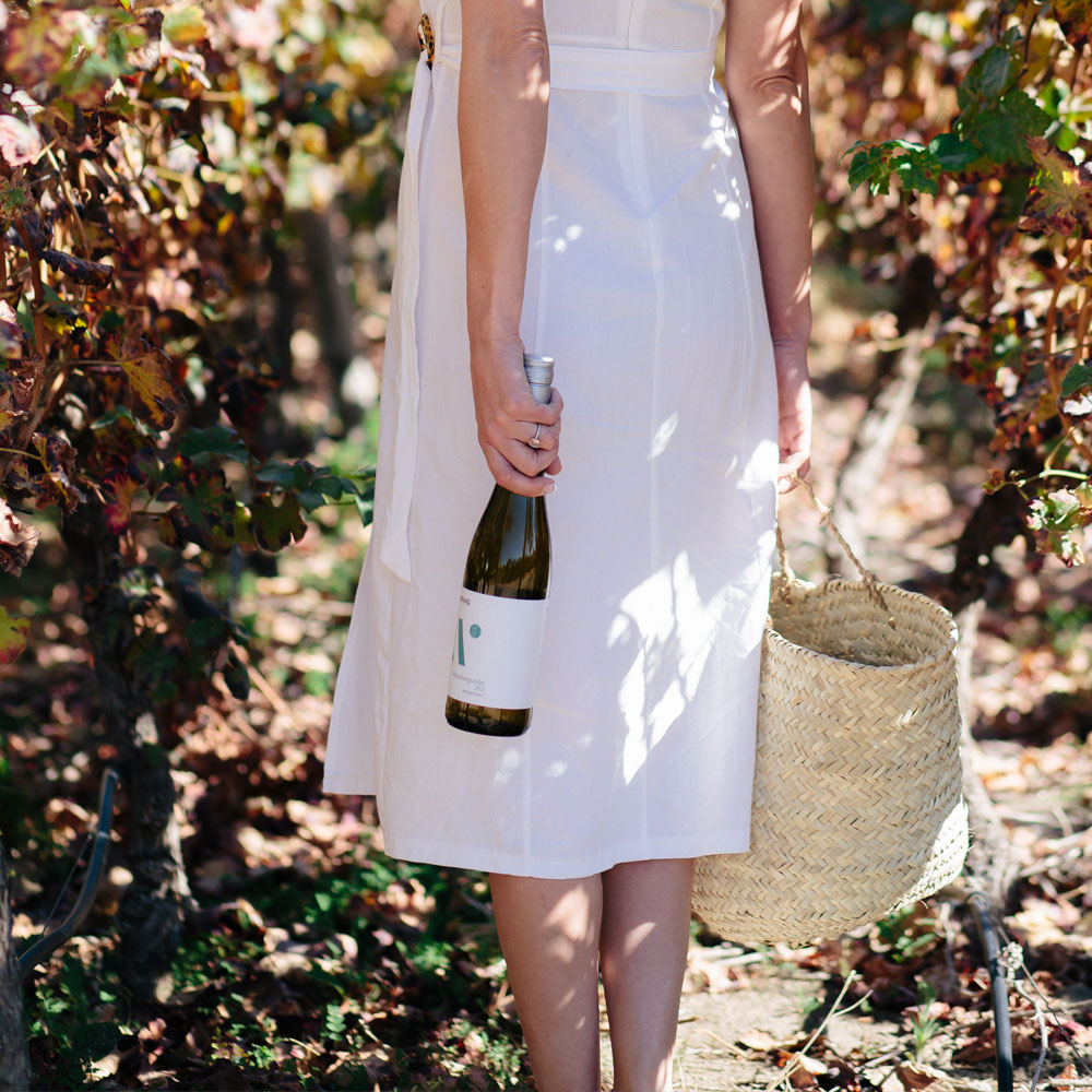 Frau in weißem Kleid trägt eine Weinflasche und einen Korb in einem Weinberg