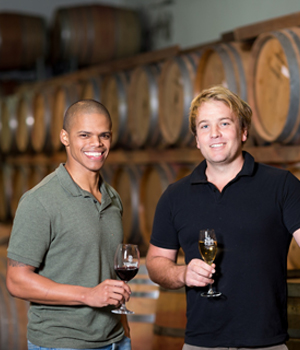 Zwei lächelnde Männer mit Weingläsern in der Hand stehen vor einer Wand mit Holzfässern in einer Weinkellerei oder einem Weinkeller.