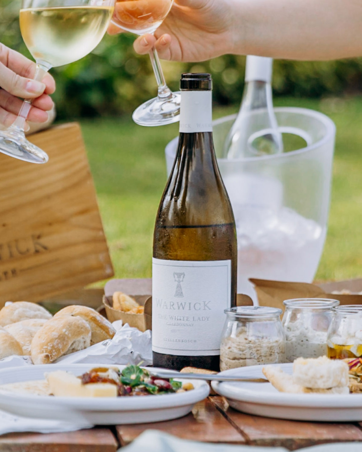 Zwei Personen stoßen mit Weingläsern an einem Picknicktisch an, der mit Brot, Käse, Aufstrichen und einer Flasche Warwick The White Lady Wein gedeckt ist, mit einem Weinkühler und einer Holzkiste im Hintergrund.