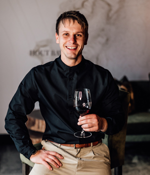 Ein junger Mann mit kurzen braunen Haaren, der ein schwarzes Hemd und eine beigefarbene Hose trägt, lächelt, während er sitzt und ein Glas Rotwein in einer Hand hält, vor einem unscharfen Innenhintergrund.