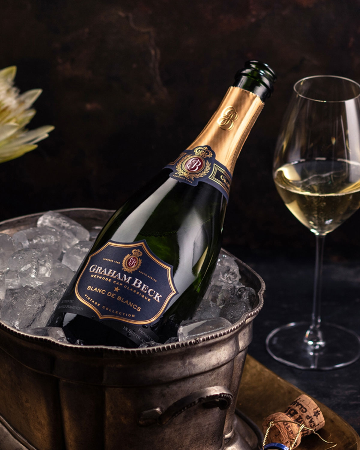 Eine Flasche Graham Beck Blanc de Blancs Sekt ruht in einem mit Eis gefüllten Metalleimer neben einem gefüllten Weinglas und einem Korken, mit einer Blume im Hintergrund.