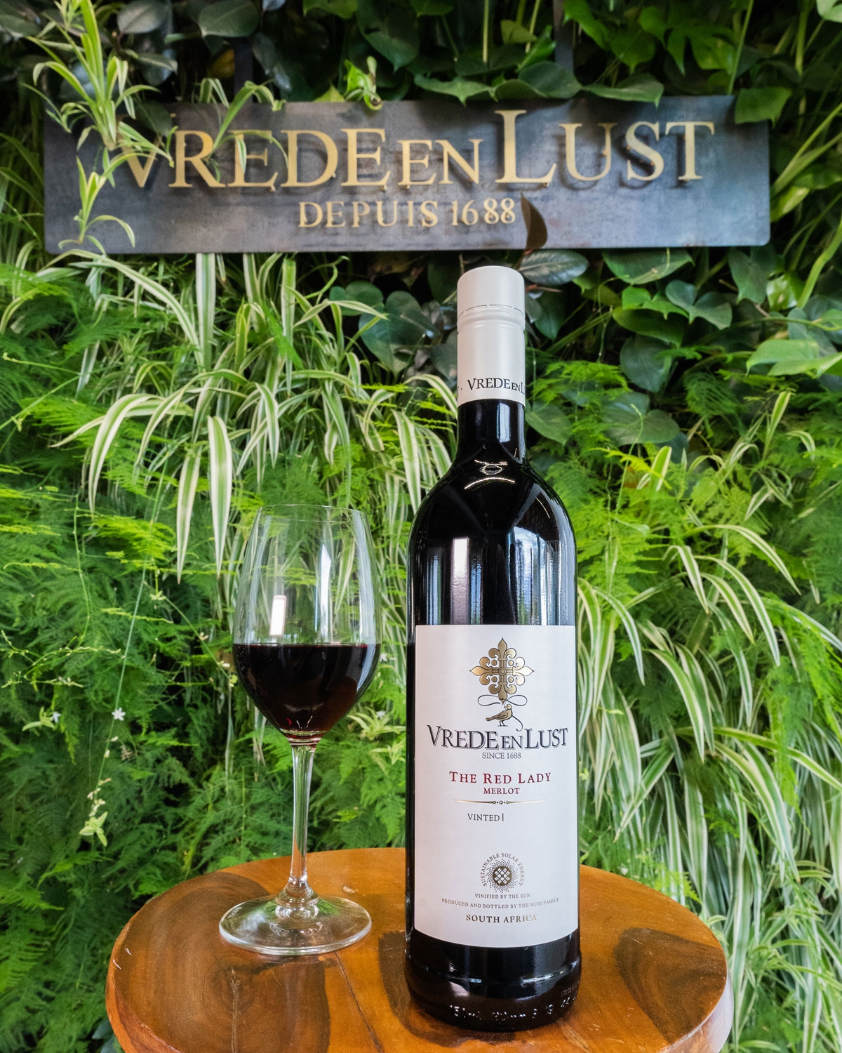 Eine Flasche Vrede en Lust Rotwein und ein gefülltes Weinglas stehen auf einem Holztisch vor einer üppigen grünen Pflanzenwand und einem Schild mit der Aufschrift Vrede en Lust Depuis 1688.