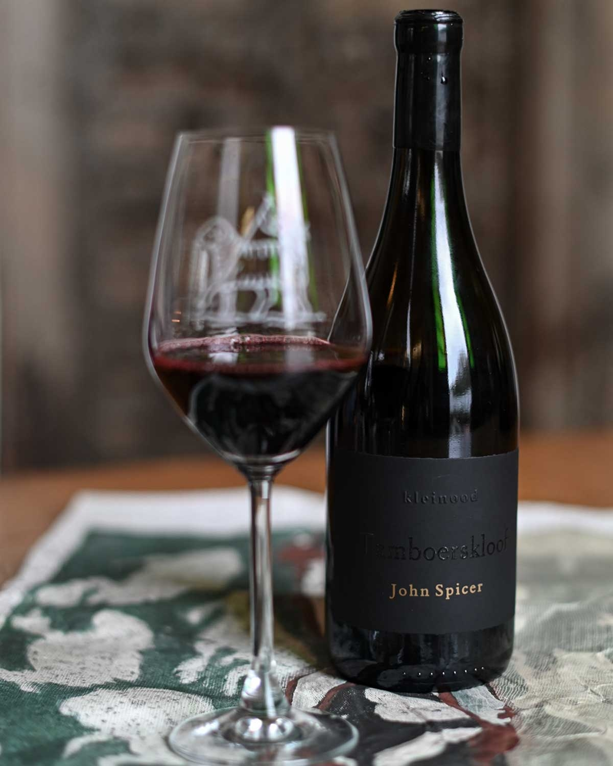 Ein Glas und eine dunkle Rotweinflasche mit der Aufschrift Tamboerskloof John Spicer stehen auf einem gemusterten Tuch, wobei der Rotwein in der Nähe des Glasbodens verschüttet wurde.