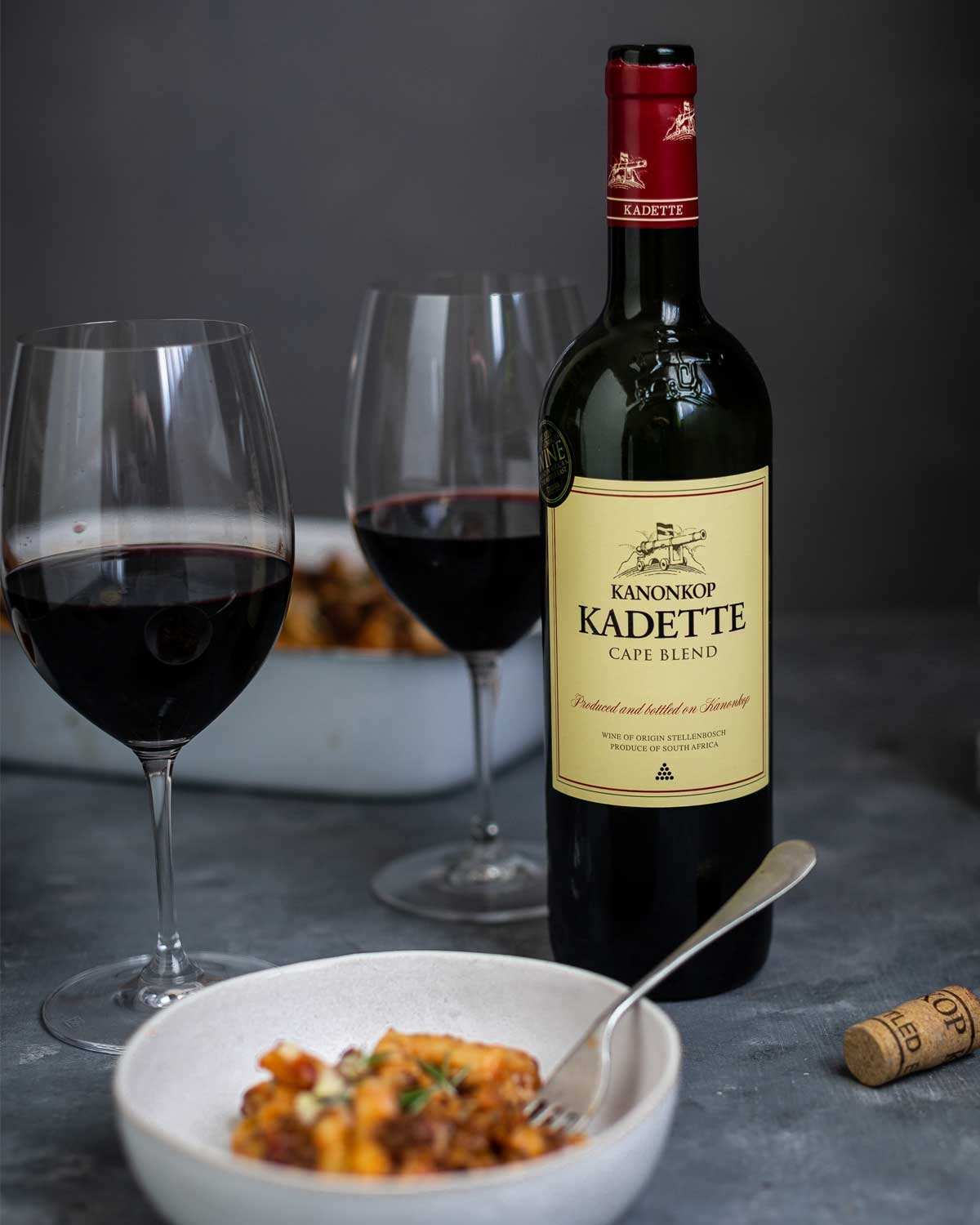 Eine Flasche Kanonkop Kadette Cape Blend Rotwein mit rotem Verschluss und cremefarbenem Etikett mit Text und der Abbildung einer Kanone. Der Wein wird in Stellenbosch, Südafrika, hergestellt.