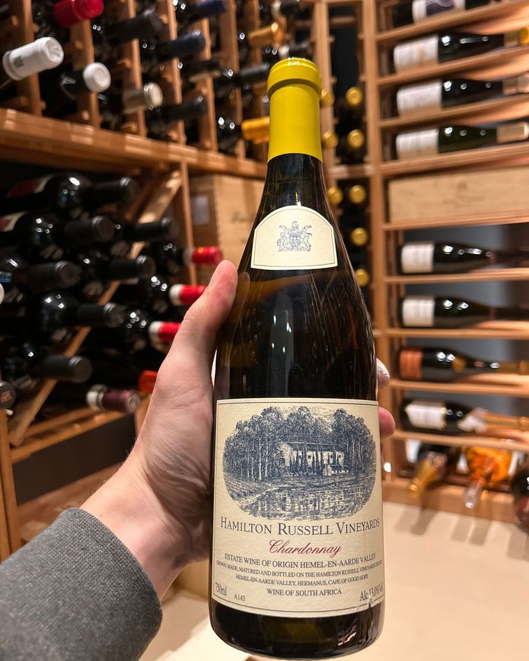 Eine Hand hält eine Flasche Hamilton Russell Vineyards Chardonnay vor einem Regal mit verschiedenen Weinflaschen in einem Weinkeller oder -laden.