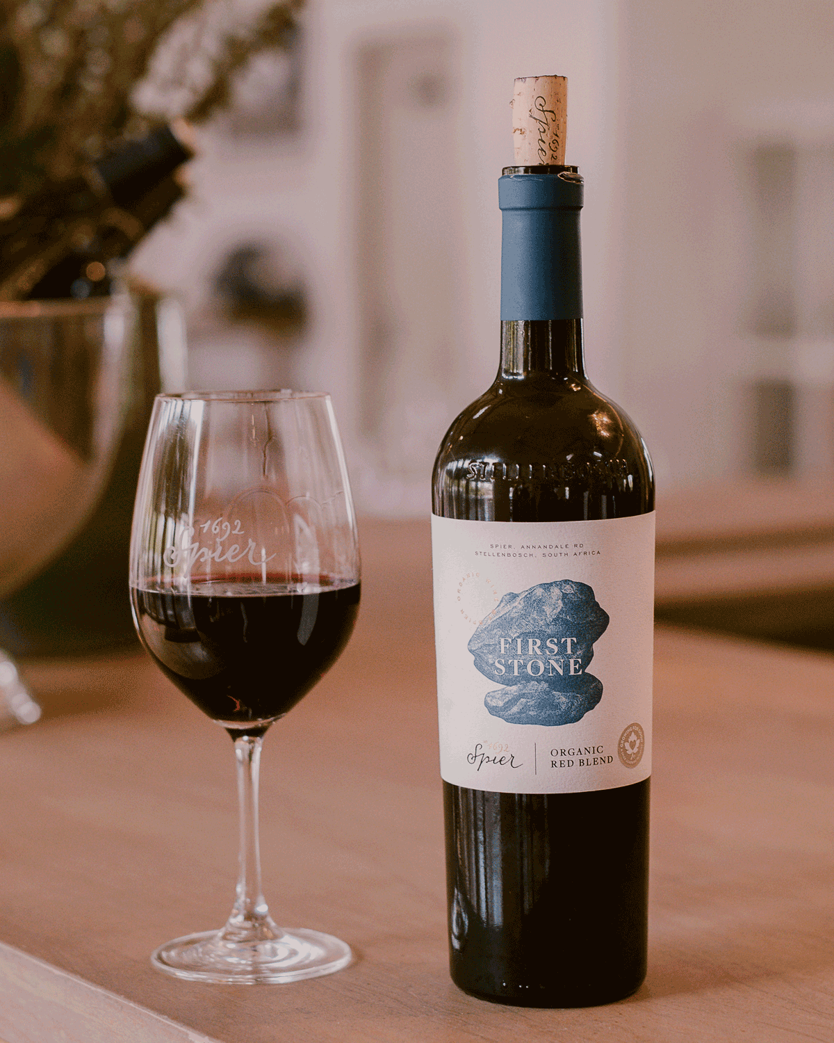 Eine Flasche First Stone Organic Red Blend Wein steht auf einem Holztisch neben einem Glas Rotwein, mit verschwommenem Hintergrunddekor und einem Weinkühler.