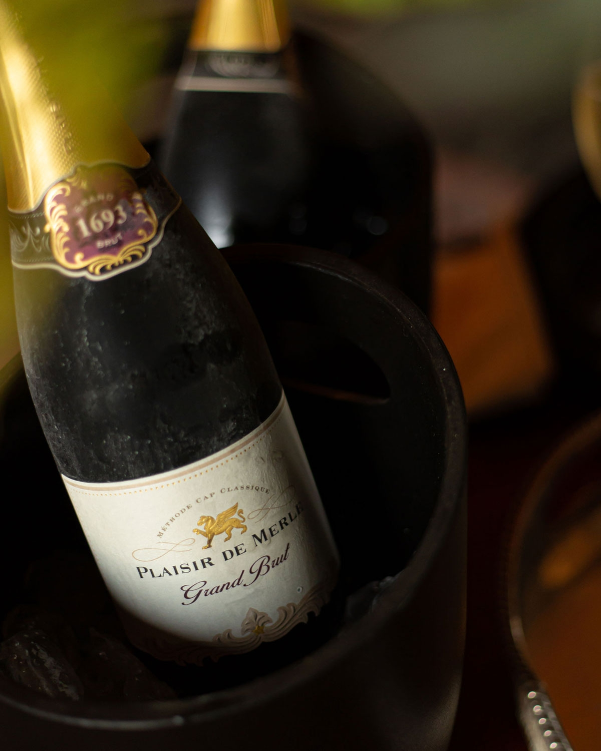 Eine Flasche Plaisir de Merle Grand Brut Sekt mit goldenem Deckel, violett-goldenem Halsetikett und weißem Hauptetikett mit elegantem Text auf einfarbigem Hintergrund.