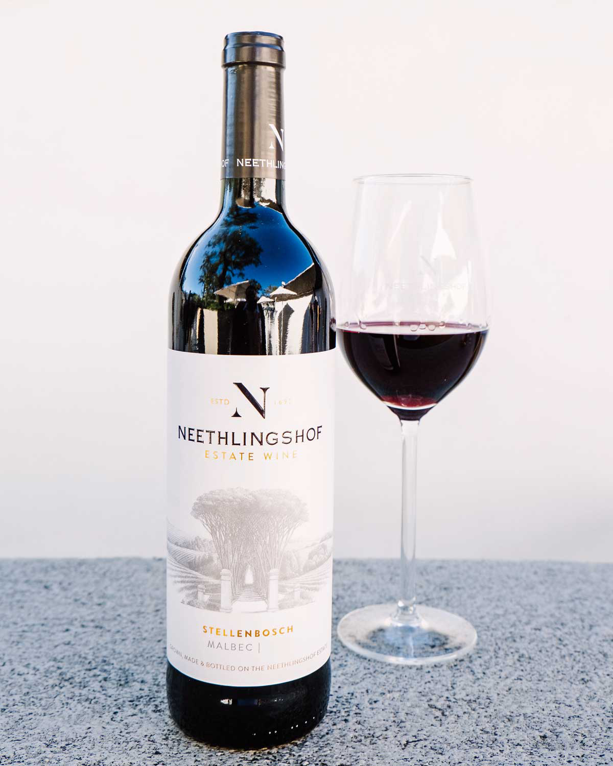 Eine Flasche Neethlingshof Estate Wine, Stellenbosch Malbec, mit einem illustrierten Etikett, das Bäume und einen Weg zeigt. Das Etikett ist cremefarben mit schwarzer und goldener Schrift.