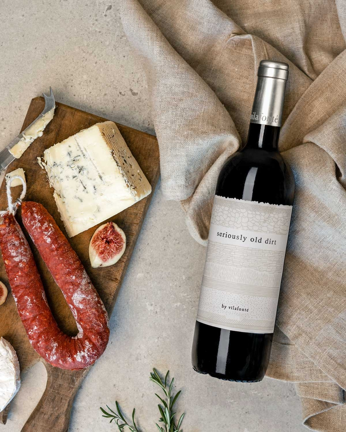 Eine Flasche Rotwein mit der Aufschrift "seriously old dirt" liegt auf einem beigen Tuch neben einem Holzbrett mit Blauschimmelkäse, Salami, Feigen und einem Zweig Rosmarin auf einer hellen Steinfläche.