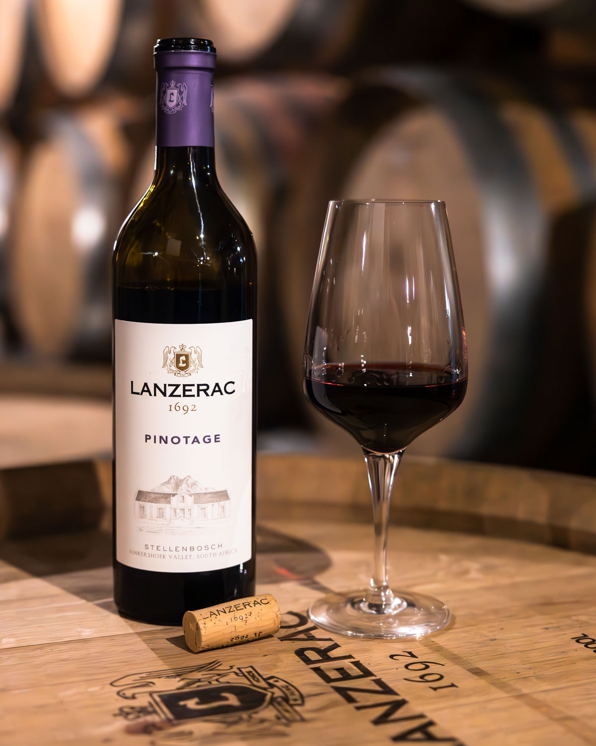 Eine Flasche Lanzerac Pinotage-Wein steht neben einem gefüllten Weinglas und einem Korken auf einem Holzfass, mit verschwommenen Weinfässern im Hintergrund.