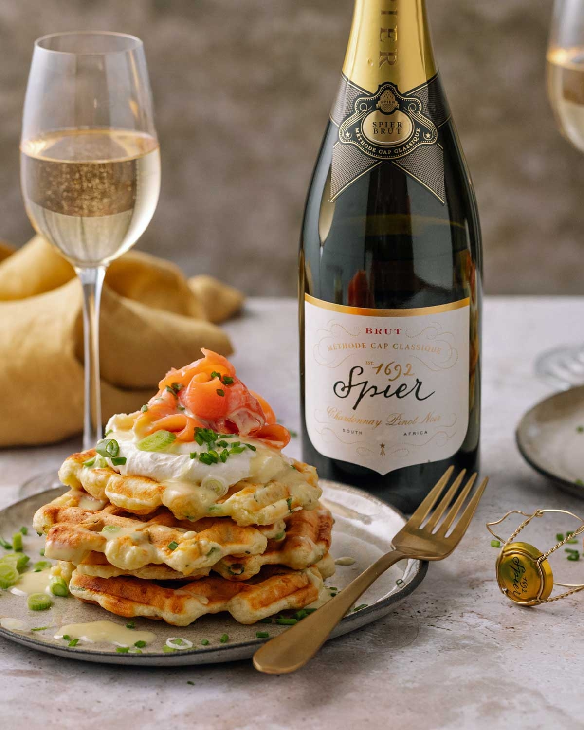 Ein Stapel herzhafter Waffeln mit Sahnesoße, Räucherlachs und Kräutern wird auf einem Teller mit einer Gabel, einem Glas Sekt und einer Flasche Spier brut Sekt serviert.