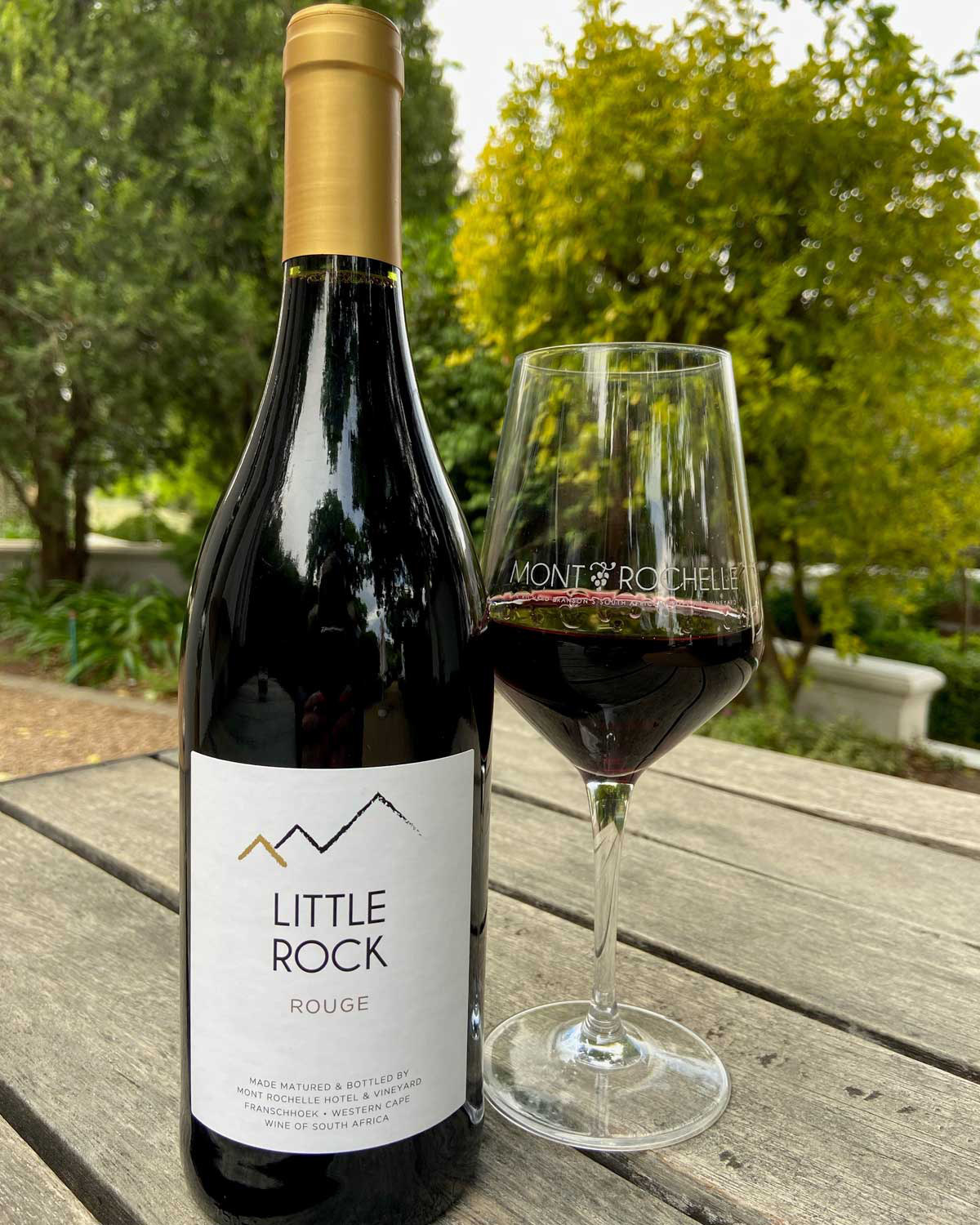 Eine Flasche des Rotweins Mont Rochelle Little Rock Rouge mit einem Deckel aus Goldfolie und einem weißen Etikett mit schwarzen Bergkonturen und goldenen Akzenten. Der Wein kommt aus Südafrika.