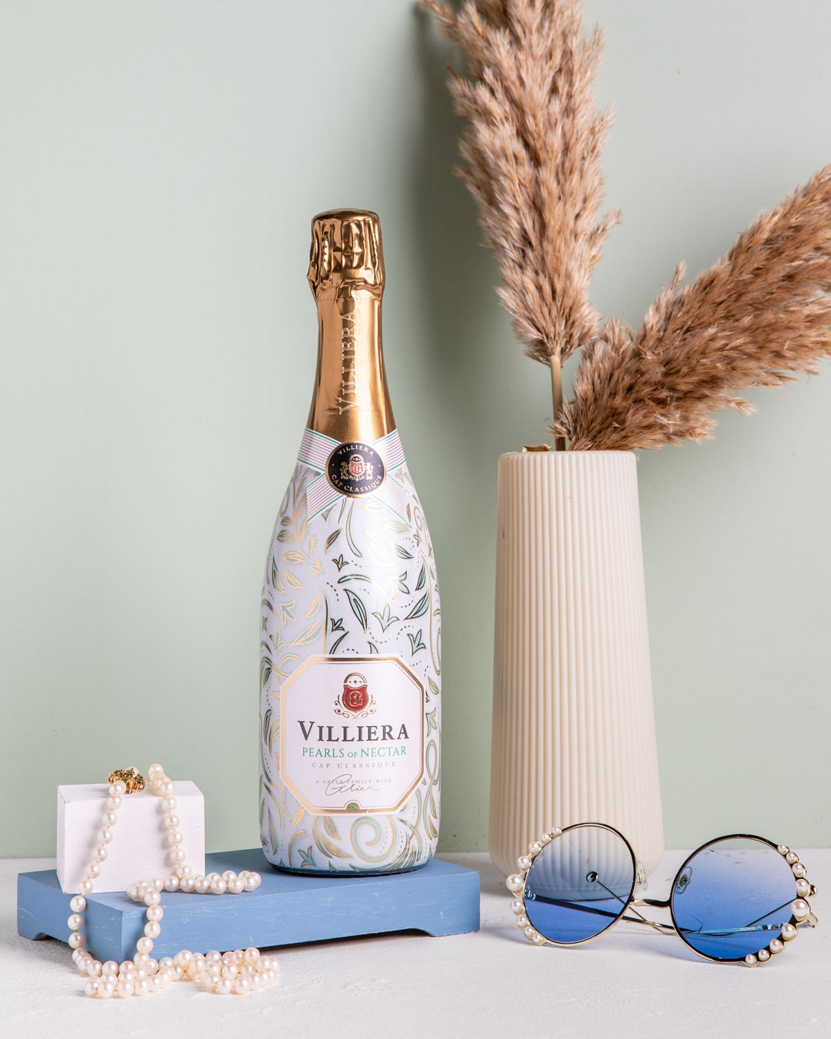 Eine Flasche Villiera-Sekt steht auf einem blauen Buch, daneben eine Vase mit getrocknetem Pampasgras, eine blau getönte Sonnenbrille und eine Perlenkette auf einem weißen Schmuckständer.
