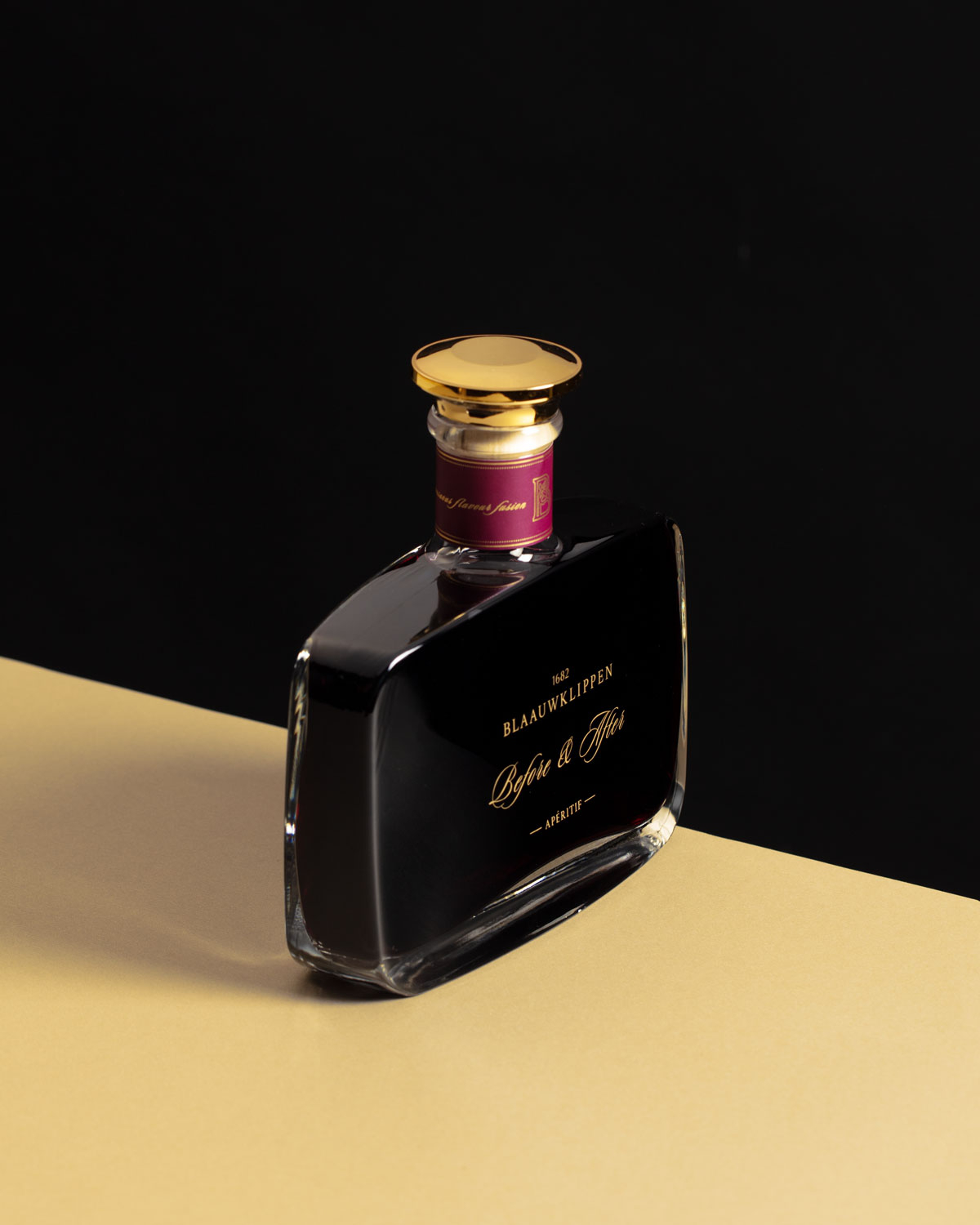 Ein stilvoller Parfümflakon aus dunklem Glas mit goldenem Verschluss und violettem Halsetikett steht auf einer beigen Fläche vor schwarzem Hintergrund. Auf dem Flakon steht ein eleganter goldener Text.