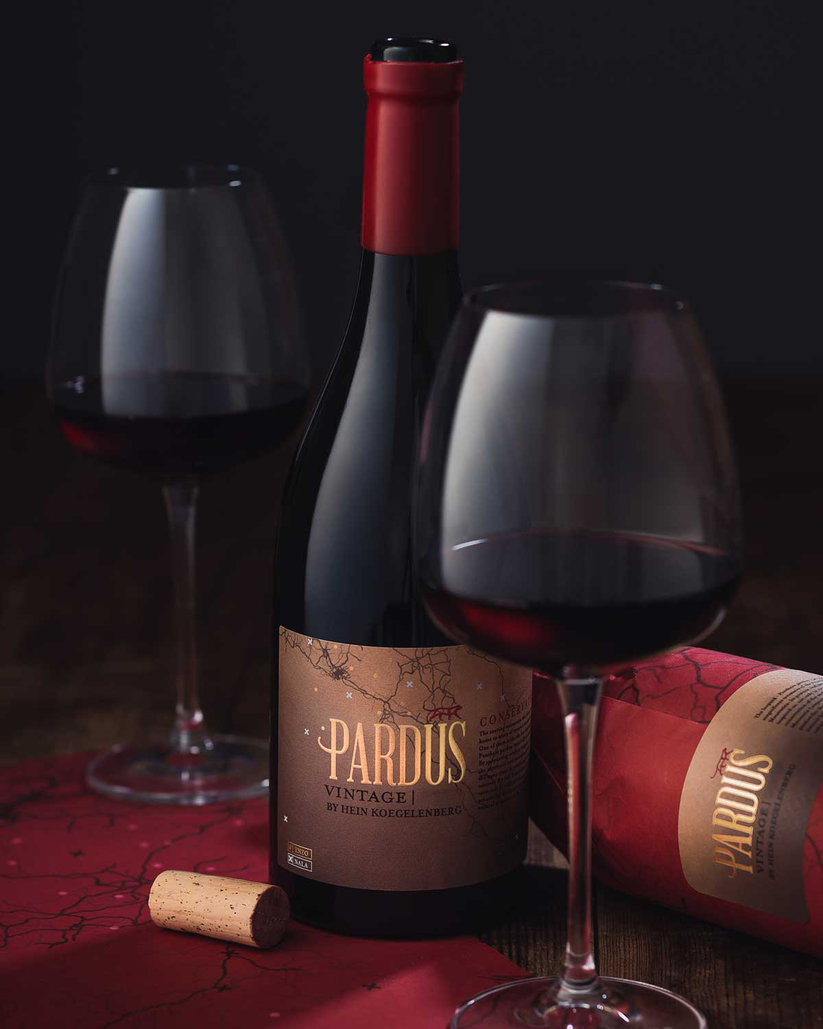 Eine Flasche Pardus Vintage Rotwein steht auf einem Holztisch mit zwei gefüllten Weingläsern, einem Korken und einer teilweise sichtbaren, in passendes Papier eingewickelten Rotweinflasche. Die Szene ist warm beleuchtet und schafft eine elegante Atmosph