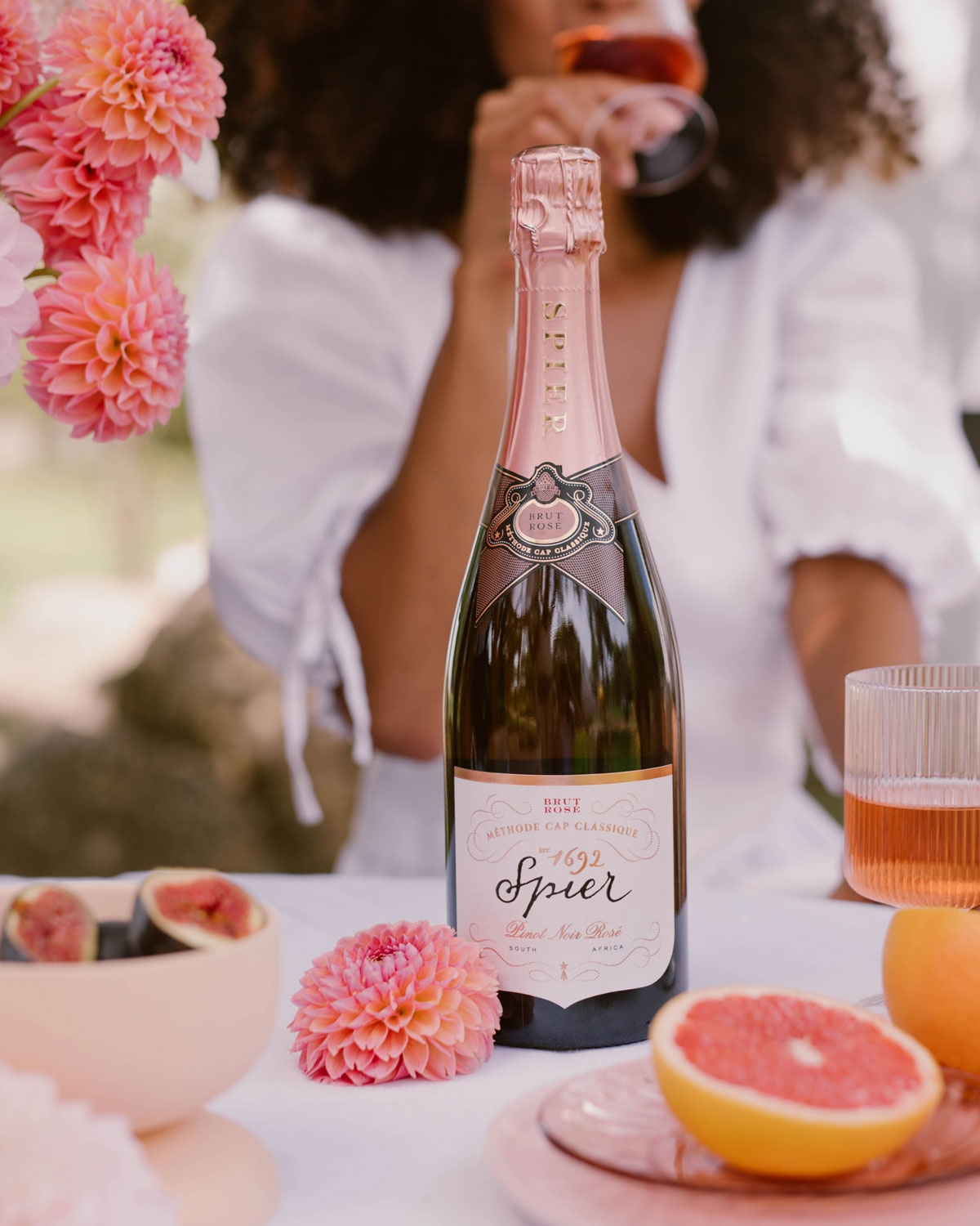 Eine Flasche Spier-Schaumwein steht auf einem Tisch mit rosa Blumen, Feigen und halbierten Grapefruits. Im Hintergrund ist eine Frau in einem weißen Kleid zu sehen, die ein Glas hält und teilweise unscharf ist.