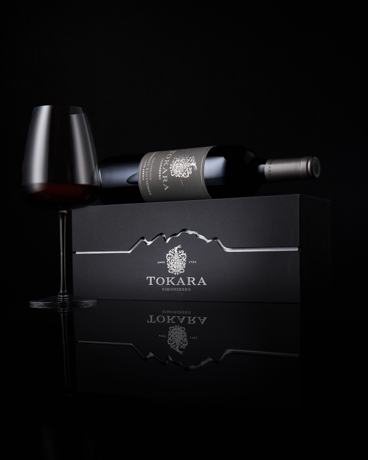 Ein Glas Rotwein steht neben einer Flasche Tokara-Wein, die auf einem schwarzen Tokara-Geschenkkarton ruht, vor einem dunklen Hintergrund, in dem sich Spiegelungen abzeichnen.