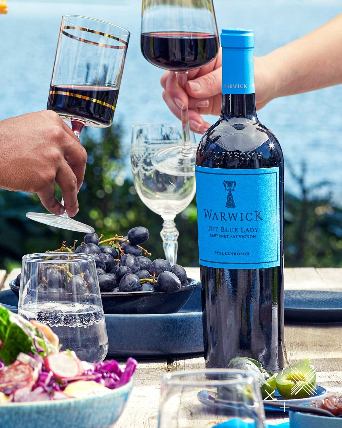 Eine Flasche Warwick The Blue Lady Cabernet Sauvignon auf einem Tisch im Freien mit Rotweingläsern, einer Schale mit Weintrauben, Salat und Wassergläsern, mit einem See und Grünpflanzen im Hintergrund.