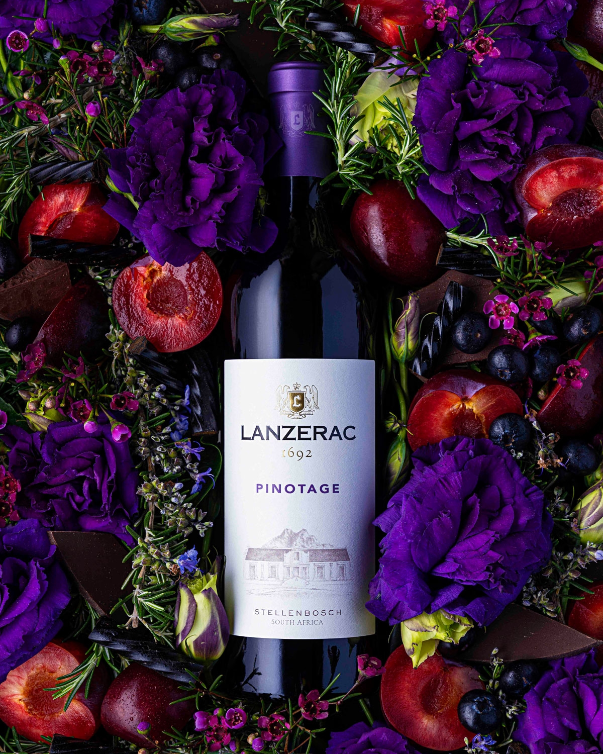 Eine Flasche Lanzerac Pinotage ist umgeben von violetten Blüten, aufgeschnittenen Pflaumen, Heidelbeeren, dunkler Schokolade, grünen Kräutern und roten Beeren.