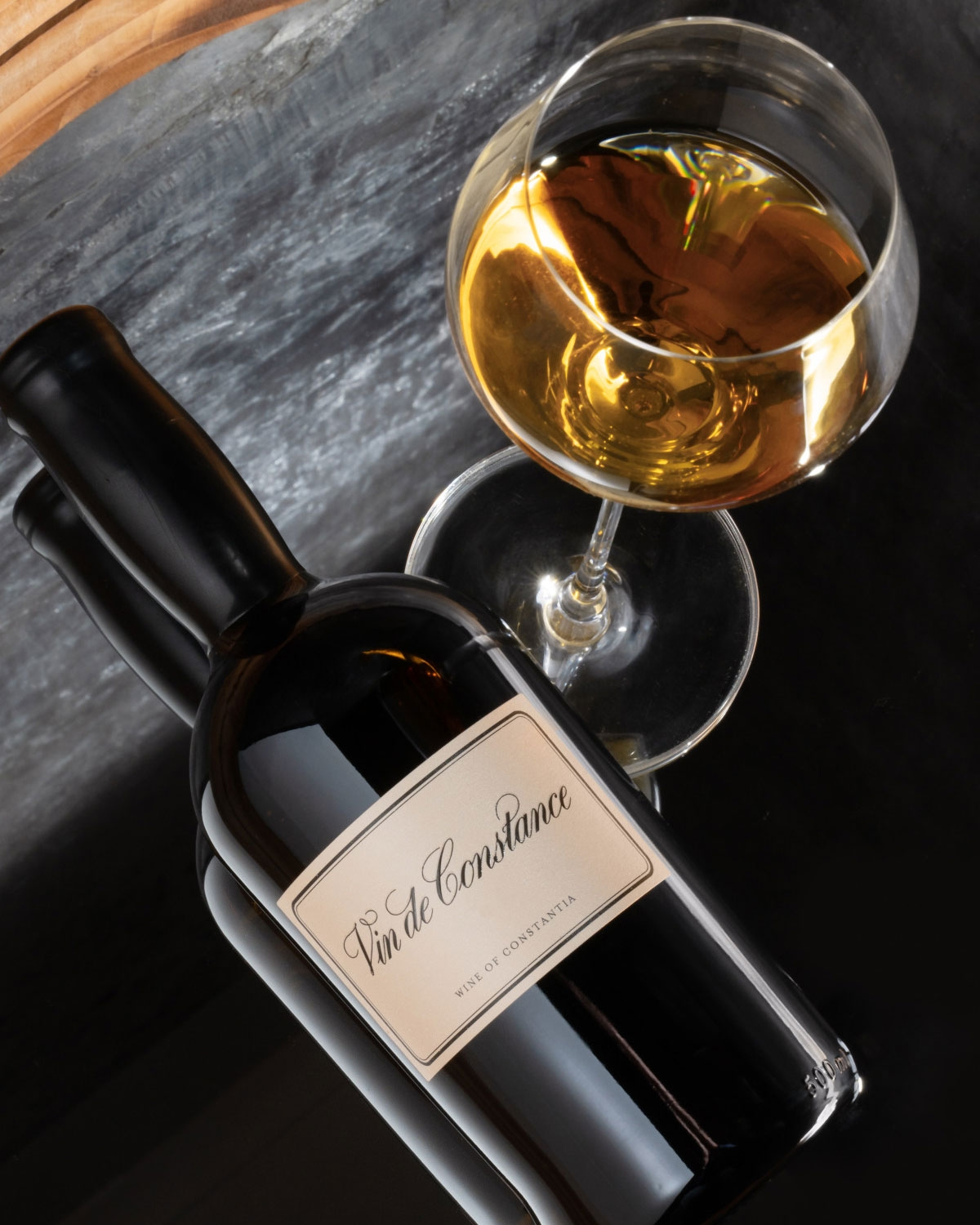 Eine Flasche Vin de Constance liegt neben einem gefüllten Weinglas auf einer dunklen Fläche, wobei der Wein eine satte goldene Farbe aufweist.