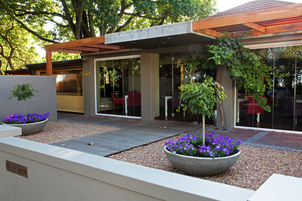Modernes Gebäude mit großen Glastüren, überhängendem Dach und lila Topfblumen in runden Pflanzgefäßen. Ein Schotterweg und ein Holzsteg führen zum Eingang, der von hohen Bäumen beschattet wird.